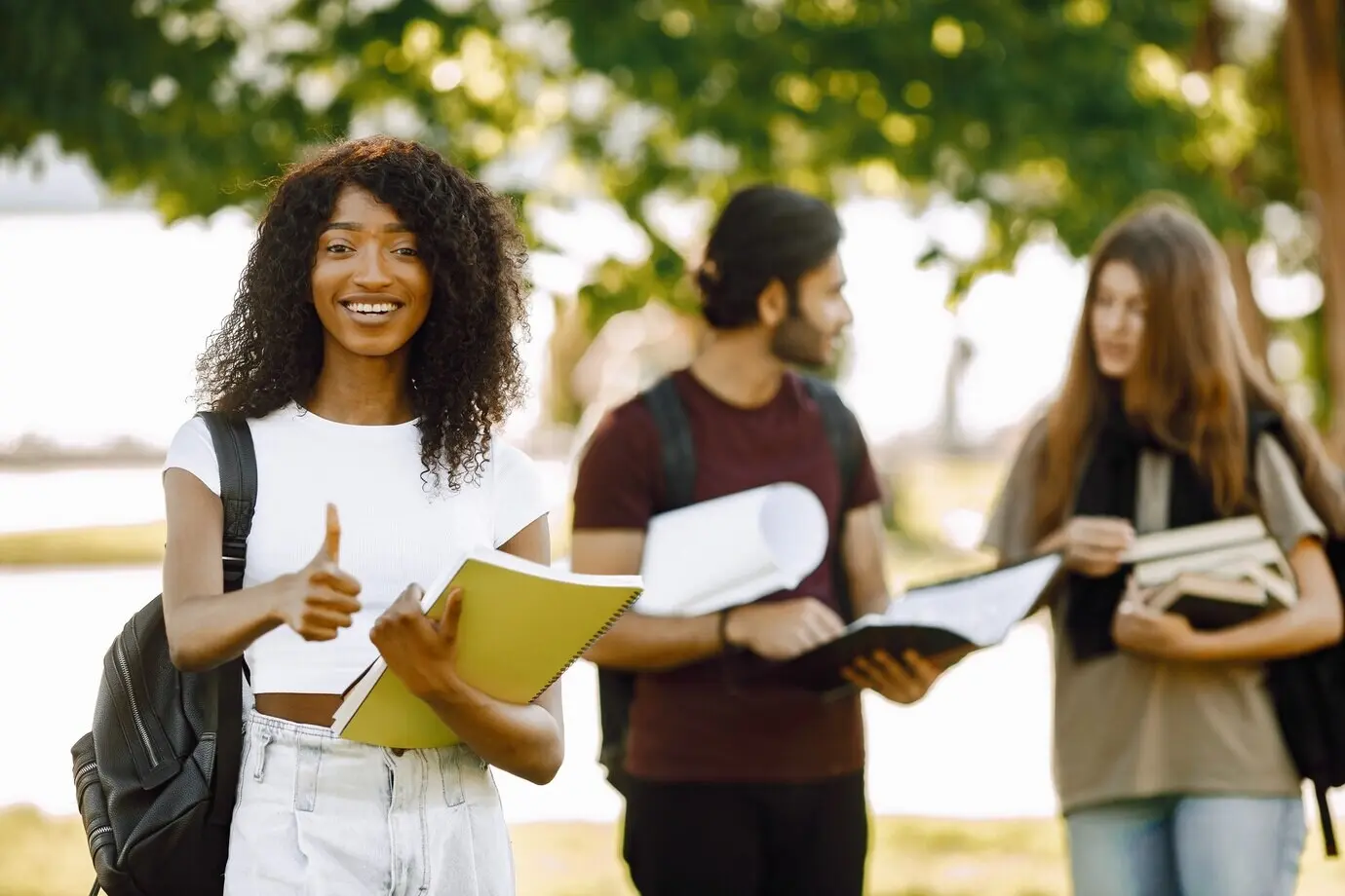 Fokus auf ein afrikanisches Mädchen, das abseits steht und den Daumen nach oben zeigt. Eine Gruppe internationaler Studierender steht zusammen im Park an der Universität.